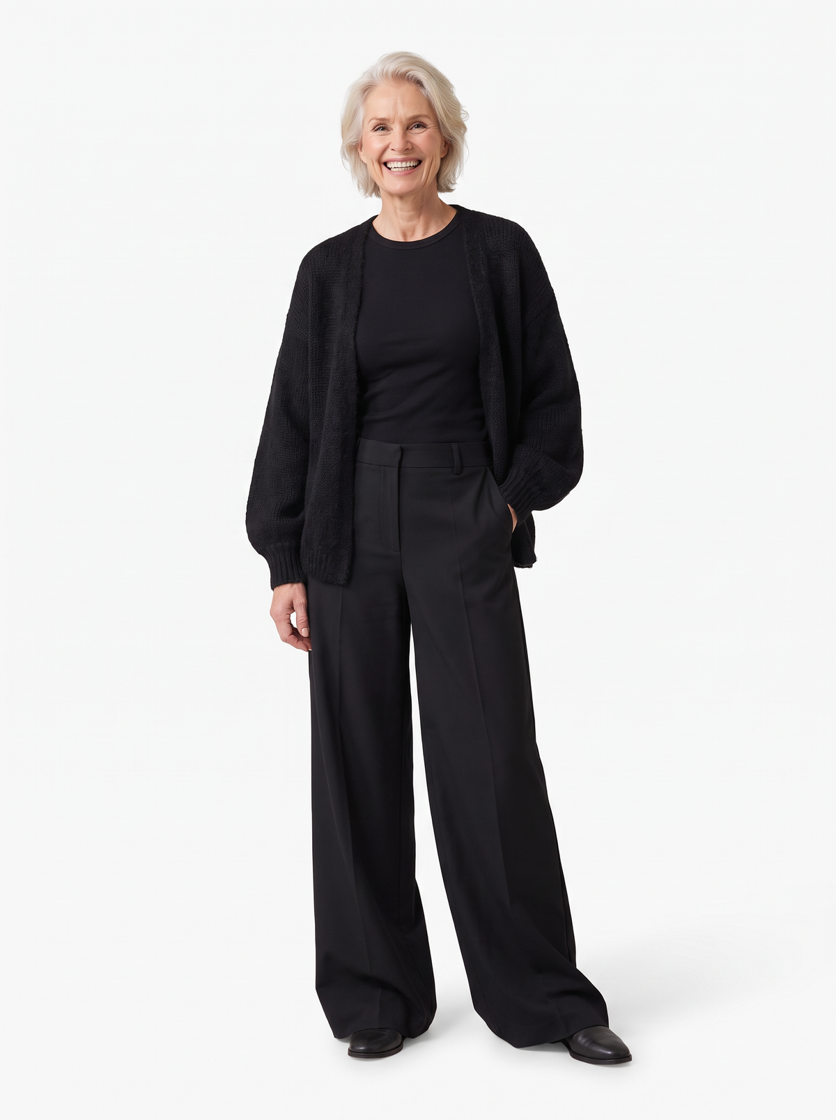 An older woman with short gray hair stands against a plain white studio background wearing a black knit cardigan over a black top and black wide-leg trousers.