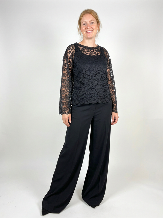 A woman modeling a black lace long-sleeve top with wide-leg black pants against a plain light gray studio background.
