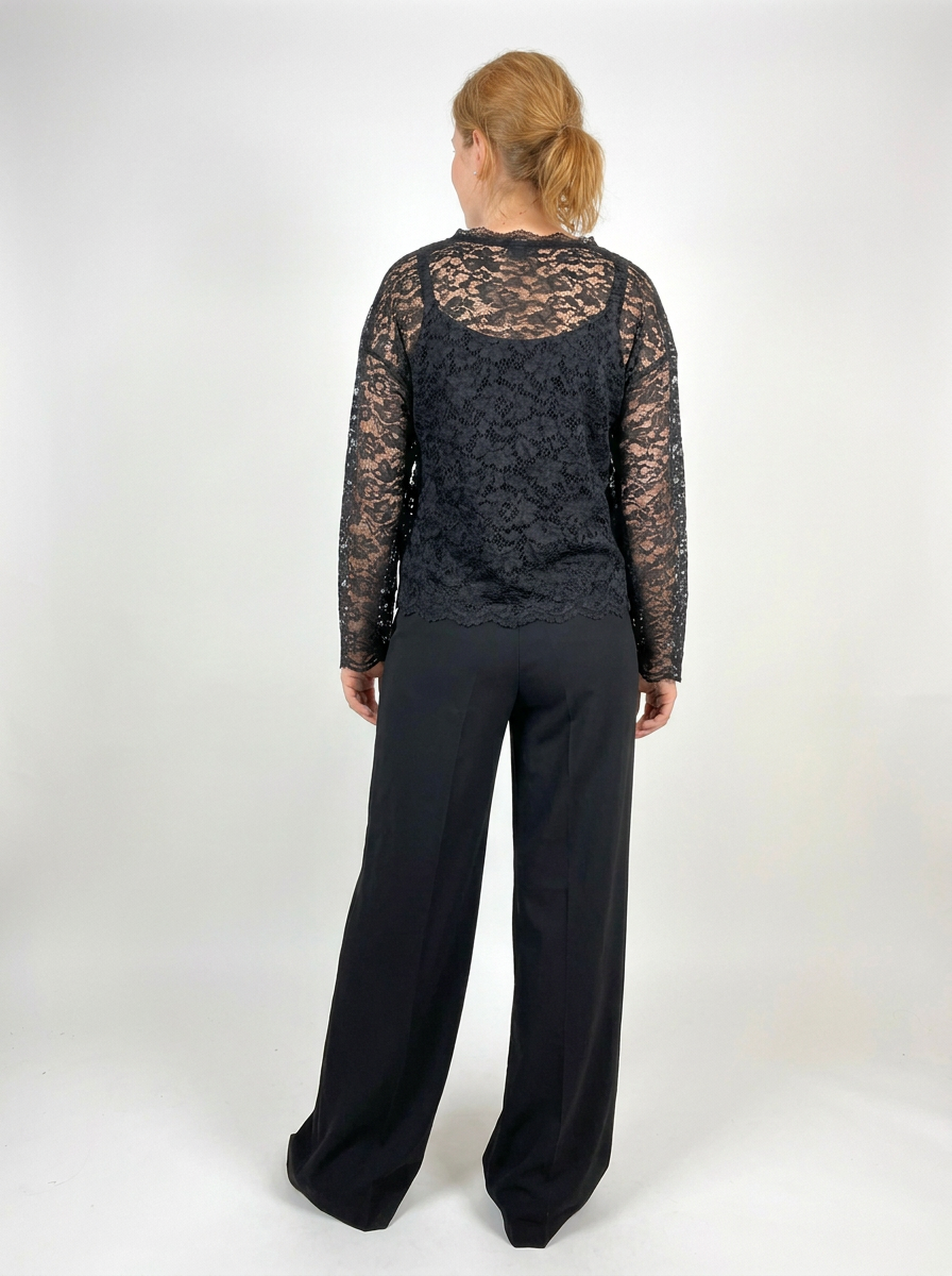 A woman viewed from the back wearing a black lace long-sleeve top and black wide-leg trousers, standing against a plain light gray studio background.