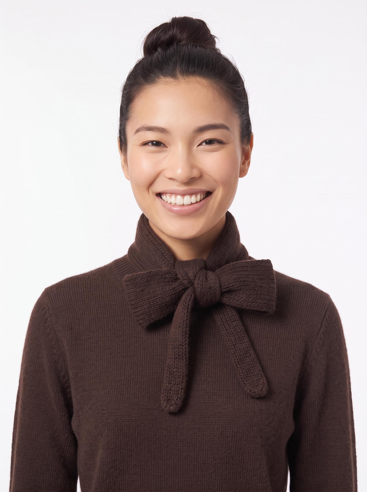 A smiling woman with her hair in a bun wearing a dark brown ribbed knit sweater with a large bow detail at the collar, set against a plain white background.