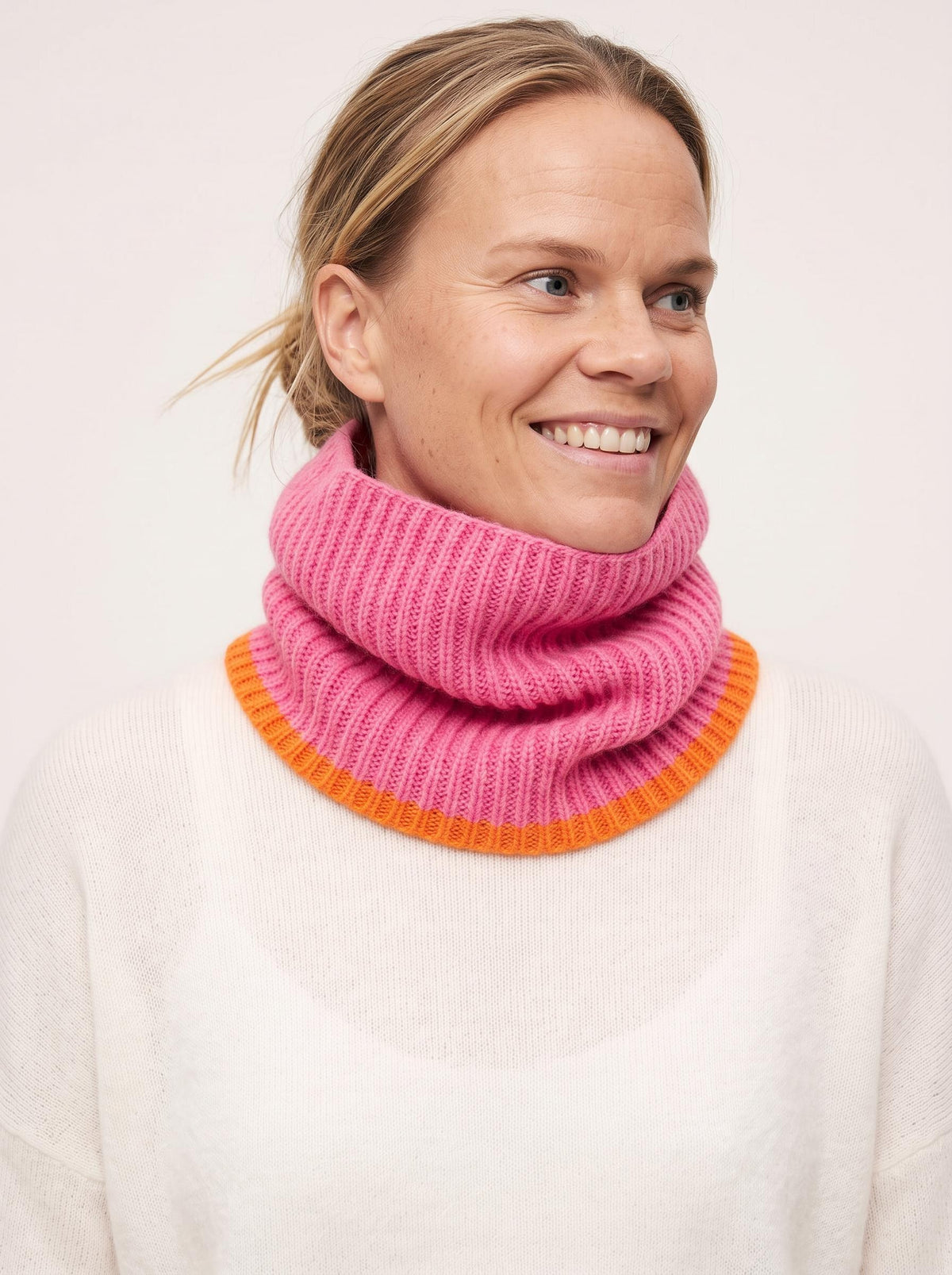 A smiling woman wearing a bright pink ribbed knit cowl neck warmer with a contrasting orange band, paired with a white sweater against a plain light background.