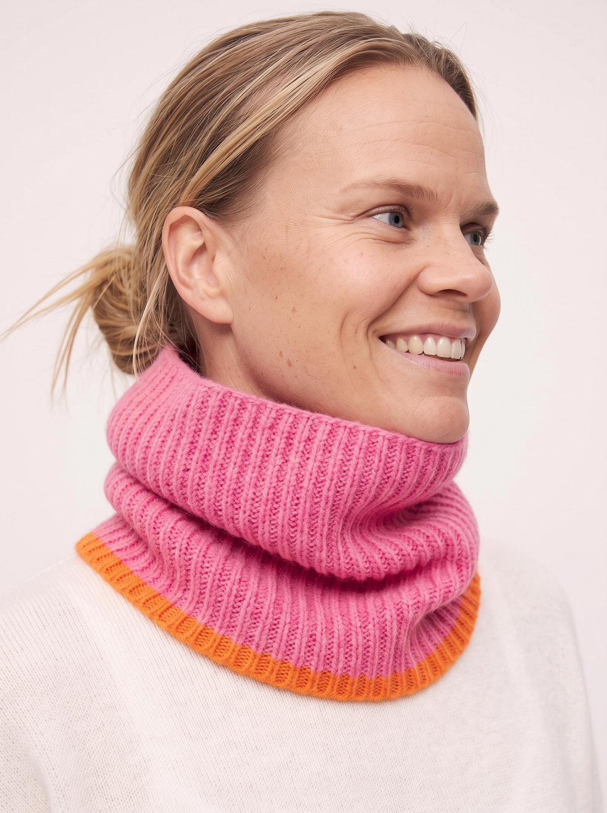 A smiling woman wearing a pink ribbed knit cowl neck warmer with a contrasting orange band at the bottom, photographed against a plain light background.